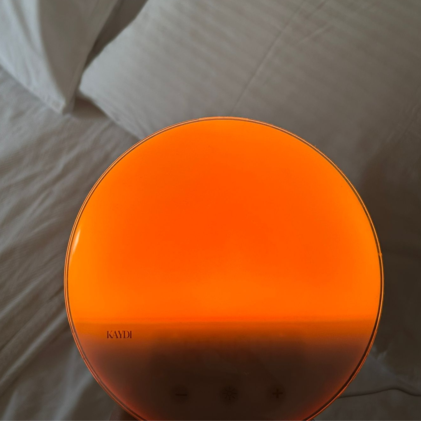 Close up of the Kaydi wake up light illuminated in warm orange light against white bedding.