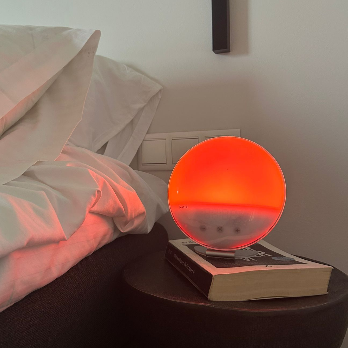 Kaydi wake up light emitting a red orange glow on a bedside table placed on top of a book next to a bed.